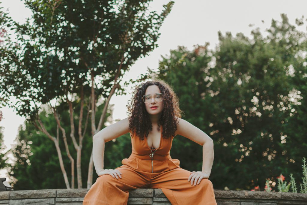 Musician Rachel Kiel crouches in a powerful stance, surrounded by trees and wearing an orange pantsuit