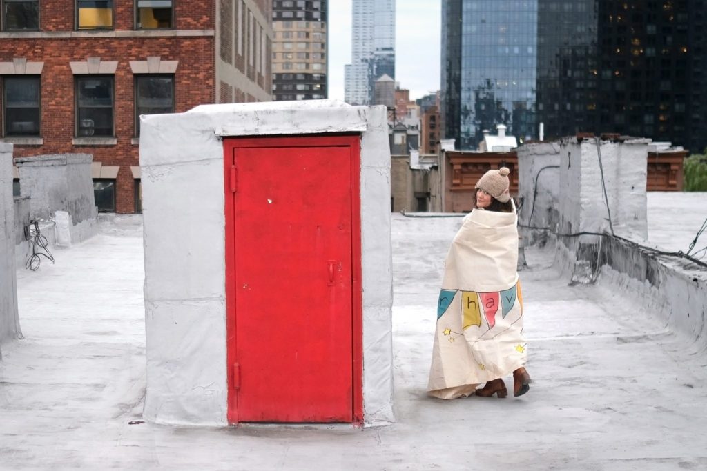 musician Tamar Haviv stands on a rooftop alongside a bright red door, wrapped in a banner that spells out her name