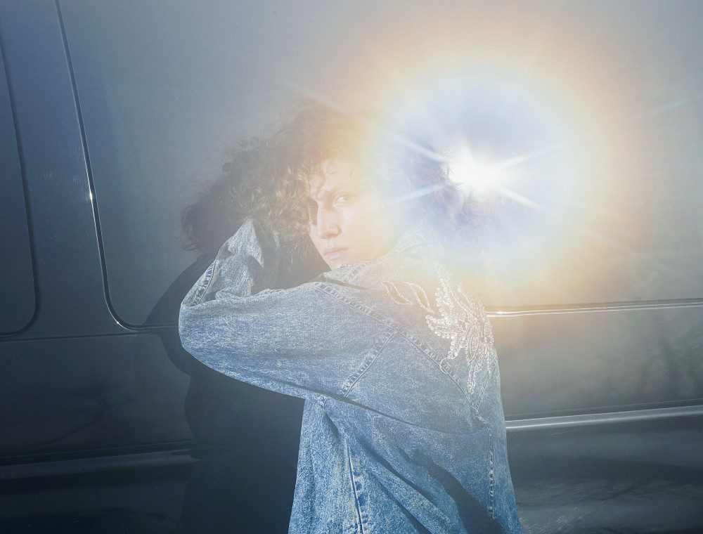 musician Squirrel Flower, wearing a denim jacket, stands partially turned away from the camera, with bright glare from the sun partially obscuring the image